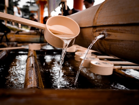 A wooden ladle is positioned under a bamboo spout with water flowing from the spout into the ladle. The arrangement is part of a traditional water purification basin, typically found at Japanese shrines. Several other ladles rest nearby, partially submerged in water.