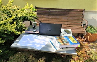 An outdoor workspace consisting of a table with a black laptop, a smartphone, a stack of books, and papers. The setting includes a wooden bench and is surrounded by greenery and potted plants. Sunlight casts soft shadows across the scene, creating a relaxed and natural atmosphere.