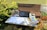 An outdoor workspace consisting of a table with a black laptop, a smartphone, a stack of books, and papers. The setting includes a wooden bench and is surrounded by greenery and potted plants. Sunlight casts soft shadows across the scene, creating a relaxed and natural atmosphere.