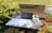 An outdoor workspace consisting of a table with a black laptop, a smartphone, a stack of books, and papers. The setting includes a wooden bench and is surrounded by greenery and potted plants. Sunlight casts soft shadows across the scene, creating a relaxed and natural atmosphere.