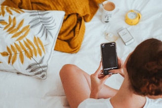 A person is seated on a bed, holding a smartphone. The bed is covered with a white sheet, a pillow with a leaf pattern, and a mustard-colored blanket. Nearby, there's a cup of coffee, an alarm clock, and a small box.