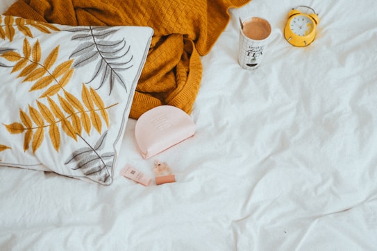 A cozy bed setting features a white pillow with yellow and gray leaf patterns, a textured mustard-colored blanket, a mug of coffee with text, a pink pouch with cosmetics, and a small yellow alarm clock on a white bedspread.