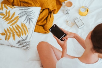 A person sits on a bed holding a smartphone. Nearby are a pillow with a leaf pattern, a mustard yellow blanket, a cup with a drink, a yellow alarm clock, a bottle of perfume, and a box. The scene suggests a calm, relaxed atmosphere.