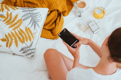 A person sits on a bed holding a smartphone. Nearby are a pillow with a leaf pattern, a mustard yellow blanket, a cup with a drink, a yellow alarm clock, a bottle of perfume, and a box. The scene suggests a calm, relaxed atmosphere.
