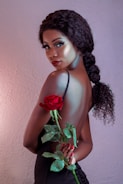A woman with curly hair in a braid poses with her back partially exposed, holding a vibrant red rose with green leaves. She has a refined and glamorous appearance, accentuated by her makeup and elegant black attire against a soft-textured background.