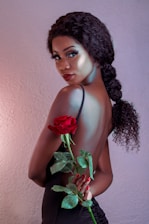 A woman with curly hair in a braid poses with her back partially exposed, holding a vibrant red rose with green leaves. She has a refined and glamorous appearance, accentuated by her makeup and elegant black attire against a soft-textured background.