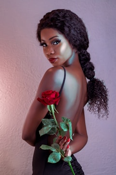 A woman with curly hair in a braid poses with her back partially exposed, holding a vibrant red rose with green leaves. She has a refined and glamorous appearance, accentuated by her makeup and elegant black attire against a soft-textured background.