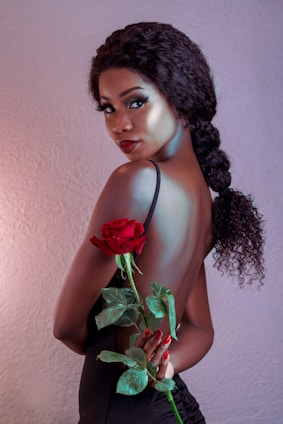 A woman with curly hair in a braid poses with her back partially exposed, holding a vibrant red rose with green leaves. She has a refined and glamorous appearance, accentuated by her makeup and elegant black attire against a soft-textured background.