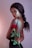 A woman with curly hair in a braid poses with her back partially exposed, holding a vibrant red rose with green leaves. She has a refined and glamorous appearance, accentuated by her makeup and elegant black attire against a soft-textured background.