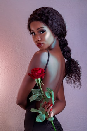 A woman with curly hair in a braid poses with her back partially exposed, holding a vibrant red rose with green leaves. She has a refined and glamorous appearance, accentuated by her makeup and elegant black attire against a soft-textured background.