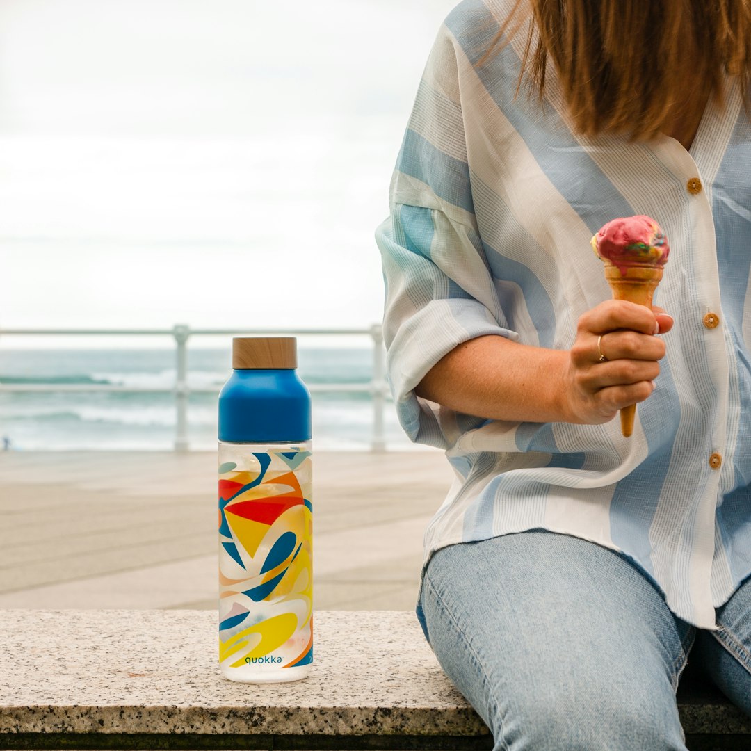 woman in white long sleeve shirt and gray denim jeans sitting on beach shore holding ice