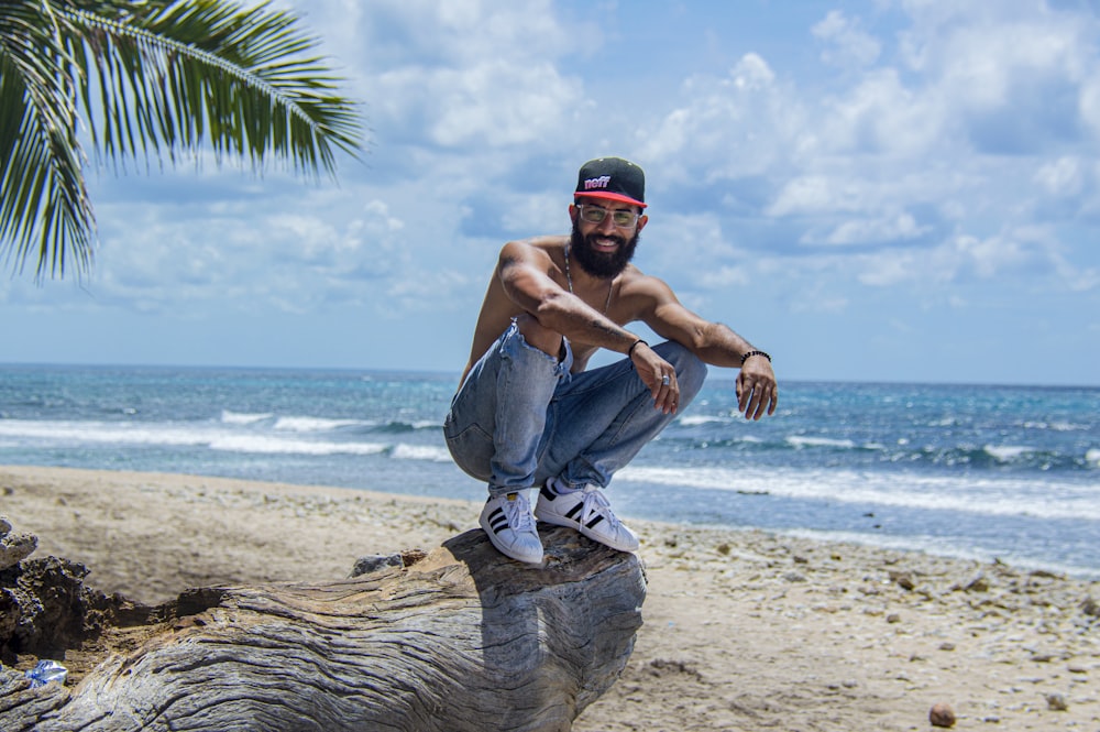 man in blue denim jeans and black cap sitting on tree trunk on beach during daytime