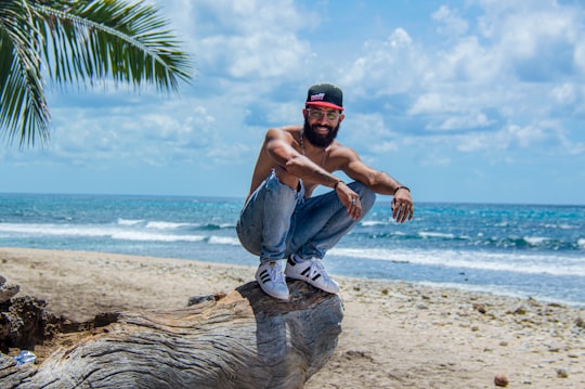 man in blue denim jeans and black cap sitting on tree trunk on beach during daytime in La Romana Dominican Republic