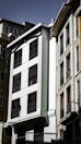 A group of multi-story residential buildings with modern architecture. The facade of the central building is predominantly white with dark windows, featuring vertical decorative elements and black wrought iron railings. Adjacent structures exhibit a mix of textures and materials, including glass and stone, under a clear sky.