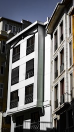 A group of multi-story residential buildings with modern architecture. The facade of the central building is predominantly white with dark windows, featuring vertical decorative elements and black wrought iron railings. Adjacent structures exhibit a mix of textures and materials, including glass and stone, under a clear sky.