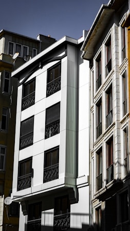 A group of multi-story residential buildings with modern architecture. The facade of the central building is predominantly white with dark windows, featuring vertical decorative elements and black wrought iron railings. Adjacent structures exhibit a mix of textures and materials, including glass and stone, under a clear sky.