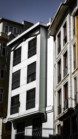 A group of multi-story residential buildings with modern architecture. The facade of the central building is predominantly white with dark windows, featuring vertical decorative elements and black wrought iron railings. Adjacent structures exhibit a mix of textures and materials, including glass and stone, under a clear sky.