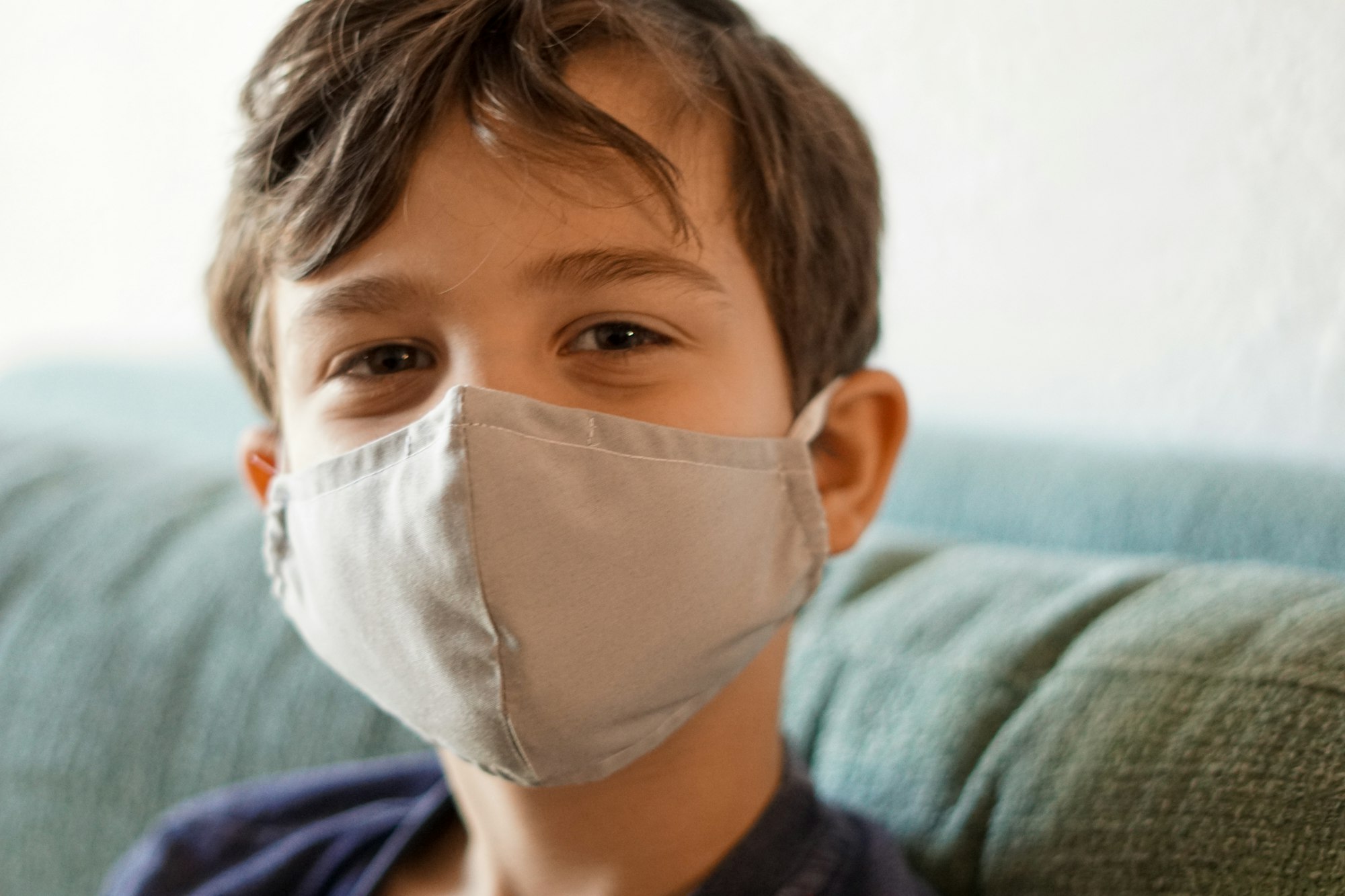 Little boy wearing a gray mask, sitting on a teal/ blue couch 