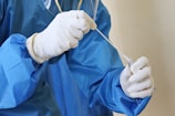 A person wearing protective blue clothing and white latex gloves is holding a swab, likely for medical testing purposes. The focus is on the hands and the swab, indicating careful handling.