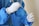 A person wearing protective blue clothing and white latex gloves is holding a swab, likely for medical testing purposes. The focus is on the hands and the swab, indicating careful handling.