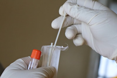 Hands wearing latex gloves are holding a swab and a specimen collection tube with an orange cap, preparing for a sample collection.