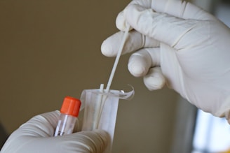 Hands wearing latex gloves are holding a swab and a specimen collection tube with an orange cap, preparing for a sample collection.