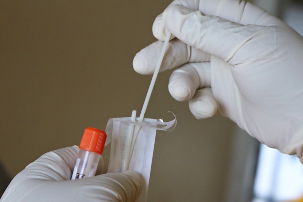 Hands wearing latex gloves are holding a swab and a specimen collection tube with an orange cap, preparing for a sample collection.
