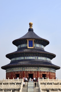 A traditional Chinese temple with a circular, tiered roof is set against a clear blue sky. Intricate designs of blue, red, and gold adorn the temple, with people visible at the entrance and on the steps leading up to it.