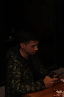 A person wearing a dark camouflage jacket is sitting at a wooden table. They are looking intently at a smartphone in their hands. The table has some scattered small items and paper.