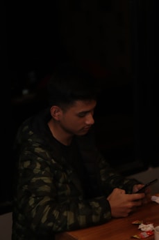 A person wearing a dark camouflage jacket is sitting at a wooden table. They are looking intently at a smartphone in their hands. The table has some scattered small items and paper.