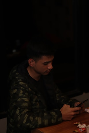 A person wearing a dark camouflage jacket is sitting at a wooden table. They are looking intently at a smartphone in their hands. The table has some scattered small items and paper.