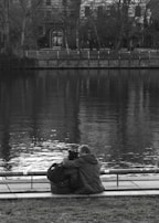 A couple sits closely together by the edge of a calm body of water, with their backs turned to the camera. They are seated on a paved area next to a park with grass in the foreground. A tree-lined embankment with a fence is visible across the water, and a large building with an arched doorway and windows stands in the background. The image is in black and white, enhancing the serene and reflective mood.