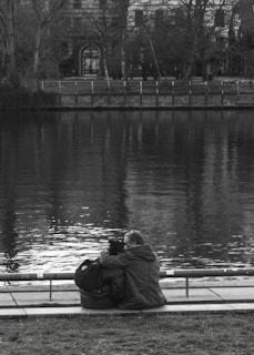 A couple sits closely together by the edge of a calm body of water, with their backs turned to the camera. They are seated on a paved area next to a park with grass in the foreground. A tree-lined embankment with a fence is visible across the water, and a large building with an arched doorway and windows stands in the background. The image is in black and white, enhancing the serene and reflective mood.