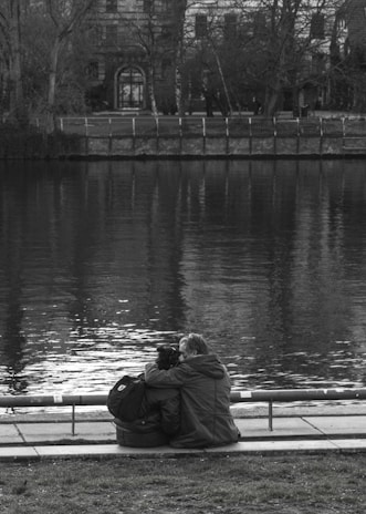 A couple sits closely together by the edge of a calm body of water, with their backs turned to the camera. They are seated on a paved area next to a park with grass in the foreground. A tree-lined embankment with a fence is visible across the water, and a large building with an arched doorway and windows stands in the background. The image is in black and white, enhancing the serene and reflective mood.