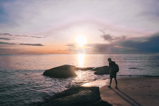 silhouette of man standing on rock near body of water during sunset