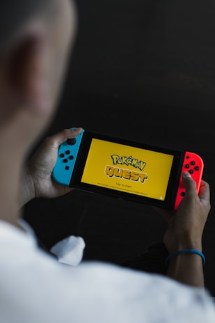 A person is holding a portable gaming console displaying the title screen for a game called 'Pokémon Quest.' The console has blue and red controllers attached to it, and the words 'Tap to start' are visible on the screen against a yellow background.