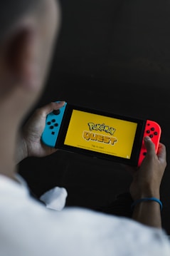 A person is holding a portable gaming console displaying the title screen for a game called 'Pokémon Quest.' The console has blue and red controllers attached to it, and the words 'Tap to start' are visible on the screen against a yellow background.