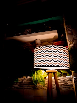 A patterned lampshade on a wooden tripod stand illuminates a room. Surrounding the lamp are decorative elements, including green leaves and a stone wall background. The design and soft lighting create a cozy and warm atmosphere.