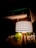 A patterned lampshade on a wooden tripod stand illuminates a room. Surrounding the lamp are decorative elements, including green leaves and a stone wall background. The design and soft lighting create a cozy and warm atmosphere.
