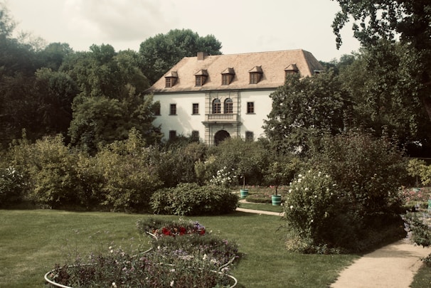 A quaint, white two-story house with a gabled roof sits amidst a lush garden. The house features several windows and a balcony on the upper floor. Surrounding the house are dense green trees and a variety of flowering plants in a well-manicured lawn. A curved path leads through the garden, enhancing the picturesque setting.