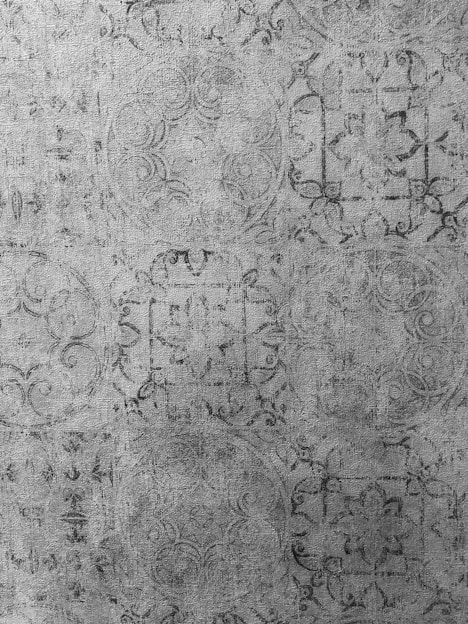Intricate, ornate patterns with swirling designs and geometric shapes appear in a monochromatic, textured style. The motifs seem to repeat in a structured yet artistic manner, resembling vintage wallpaper or fabric.