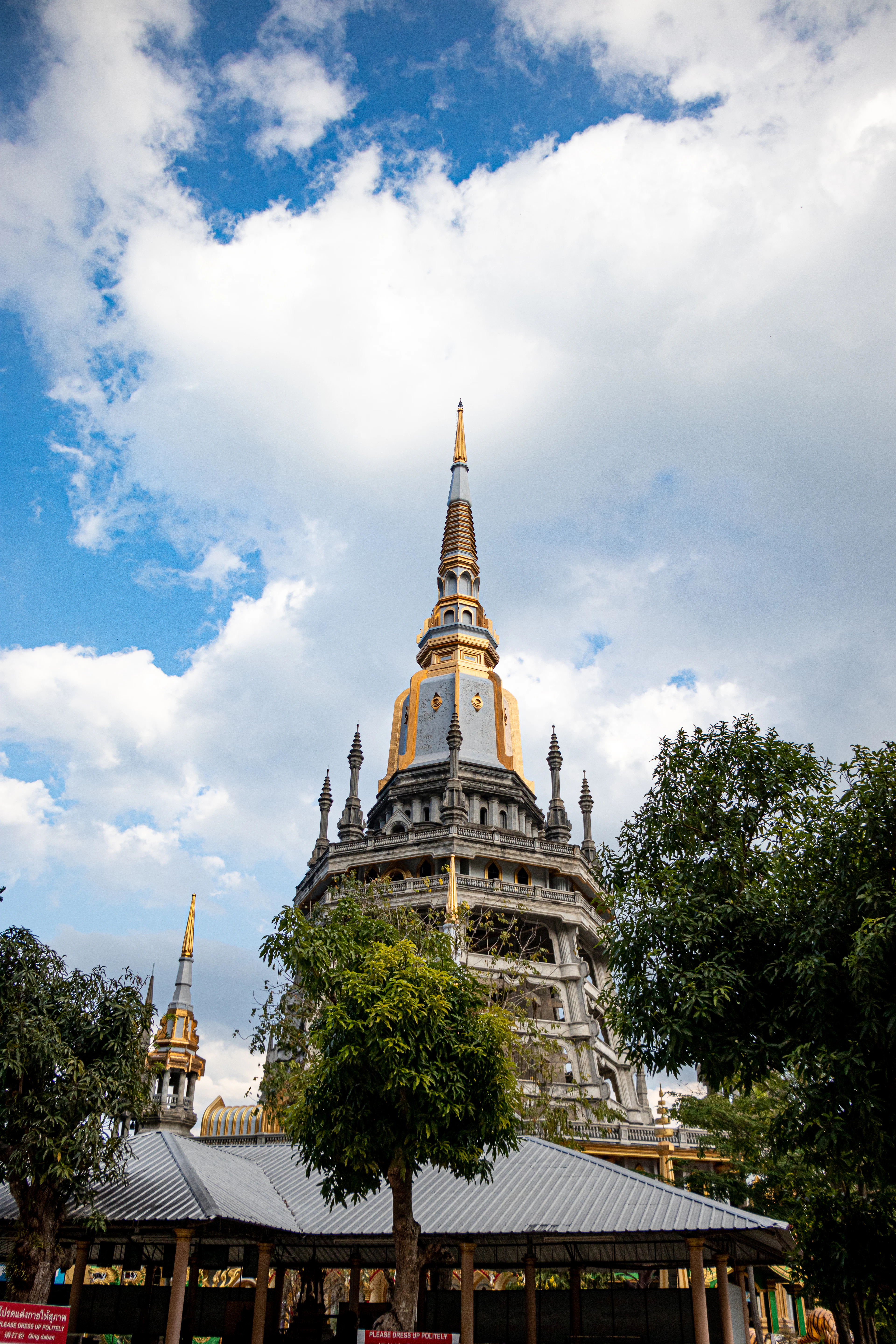 The white and gold pagoda of the Tiger Cave Temple in Krabi seen from the courtyard below, its gilded spire set against towering cumulus, mango trees in the foreground.