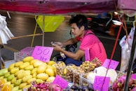A street vendor is sitting at a small fruit stall, surrounded by colorful fruits such as mangoes, dragon fruits, and longans. The vendor is wearing a pink shirt and appears to be checking a mobile phone. Handwritten price tags are displayed prominently on the fruits.