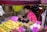 A street vendor is sitting at a small fruit stall, surrounded by colorful fruits such as mangoes, dragon fruits, and longans. The vendor is wearing a pink shirt and appears to be checking a mobile phone. Handwritten price tags are displayed prominently on the fruits.