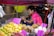 A street vendor is sitting at a small fruit stall, surrounded by colorful fruits such as mangoes, dragon fruits, and longans. The vendor is wearing a pink shirt and appears to be checking a mobile phone. Handwritten price tags are displayed prominently on the fruits.