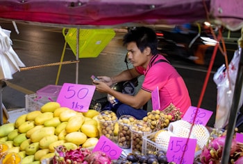 A street vendor is sitting at a small fruit stall, surrounded by colorful fruits such as mangoes, dragon fruits, and longans. The vendor is wearing a pink shirt and appears to be checking a mobile phone. Handwritten price tags are displayed prominently on the fruits.