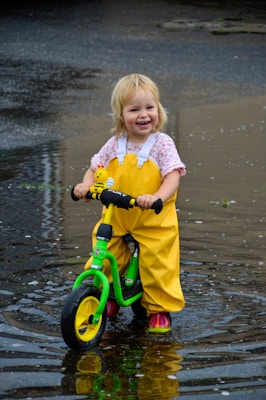 A cheerful child wearing bright yellow rain pants and floral shirt is riding a green and yellow balance bike through a shallow puddle. The child appears delighted and the water reflects part of the surroundings.