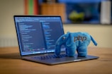 A laptop is displaying a programming code editor with multiple lines of colorful code. Beside the trackpad, there is a blue plush toy shaped like an elephant with the text 'php' embroidered on its side. The background is slightly blurred, showing hints of a cozy indoor environment.