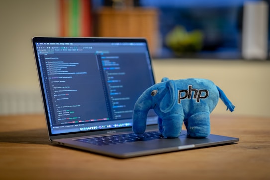 A laptop is displaying a programming code editor with multiple lines of colorful code. Beside the trackpad, there is a blue plush toy shaped like an elephant with the text 'php' embroidered on its side. The background is slightly blurred, showing hints of a cozy indoor environment.