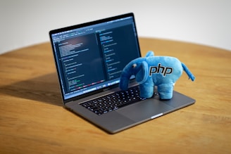A laptop displaying code on its screen sits on a wooden table, with a blue plush toy elephant branded with 'PHP' perched on its keyboard.